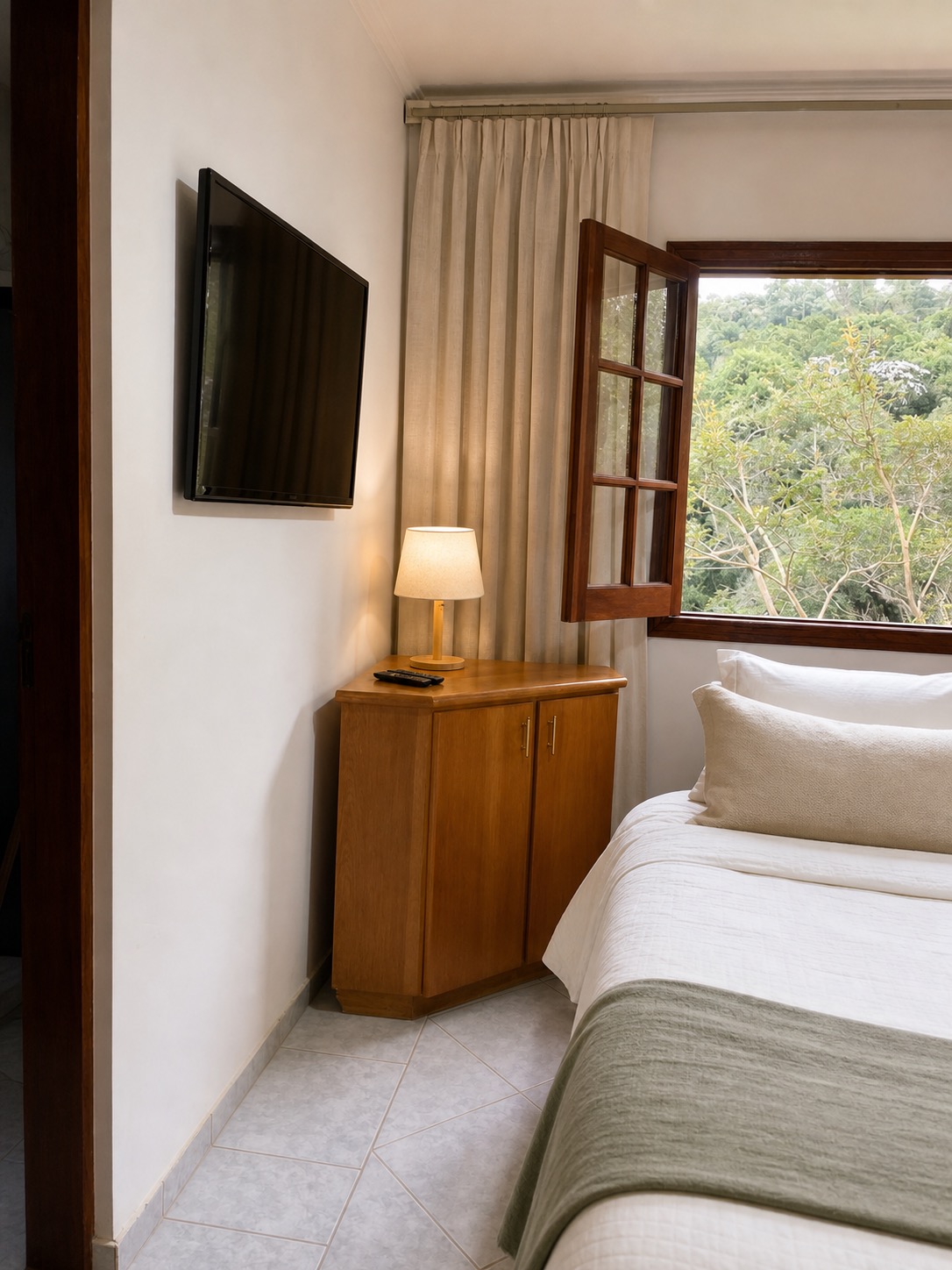 Canto de quarto com aparador de madeira, abajur acesso, TV e janela de madeira aberta para a mata